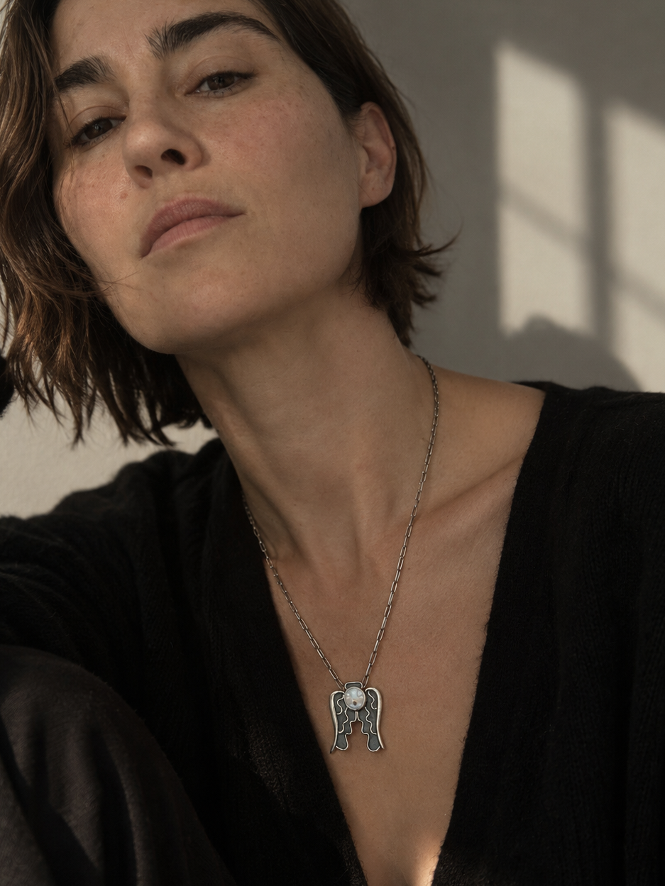 A close-up lifestyle shot of a woman with short brown hair wearing a silver winged pendant necklace and a black knit top, set against a warm-toned wall with soft window shadows.